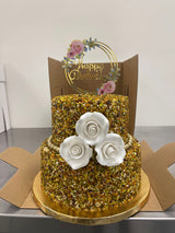 A Two-Tier Pistachio San Rival cake, covered in pistachio nuts and decorated with three frosted white roses, gold beading, and a golden birthday topper that says, "Happy Birthday."