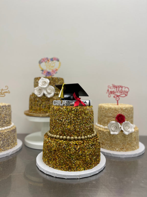 A Two-Tier Pistachio San Rival cake, covered in pistachio nuts and decorated with gold beading and a graduation cake topper. In the background, two more Two-Tier San Rival cakes are displayed. 
