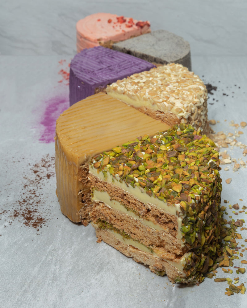 Six slices of different San Rival cakes, vertically placed: Pistachio, Coffee, Cashew, Ube, Cookie n Cream, and Strawberry. 