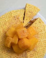 Close-up shot of a yellow Mango San Rival round cake, with one slice cut out, topped with frozen squared mango bites and crushed graham.