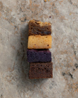 Four Overload Bars stacked on top of each other. Top to bottom: Food For The Gods, Caramel, Ube, and Chocolate. Surrounding them are scattered blurred bars. 