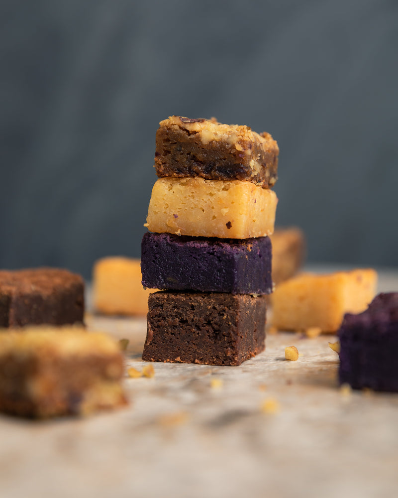 Four Overload Bars stacked on top of each other. Top to bottom: Food For The Gods, Caramel, Ube, and Chocolate. Surrounding them are scattered blurred bars. 